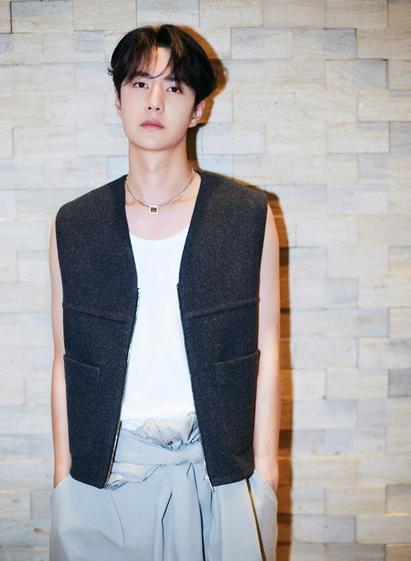 Wang Yibo standing in front of a brick wall, wearing a grey felt vest and a sleeveless white top.