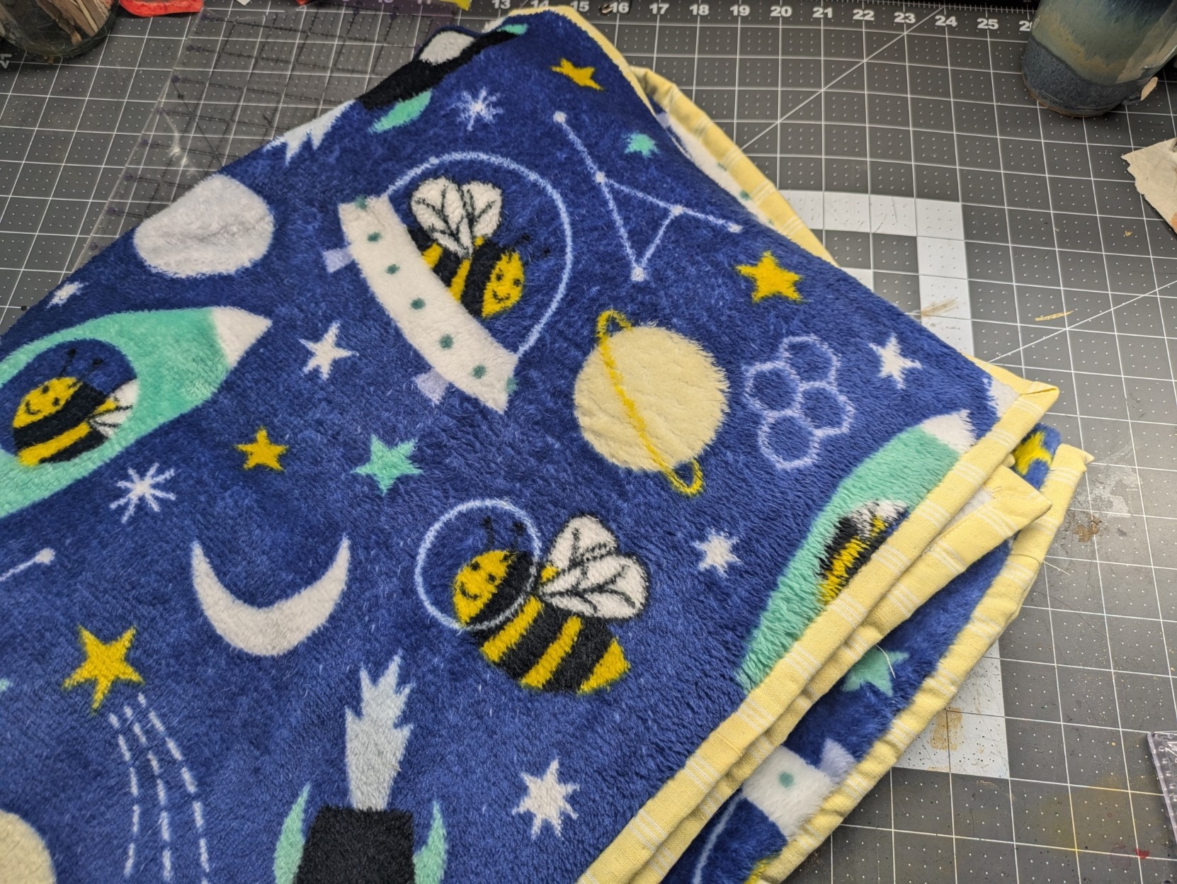 A folded blanket made of blue fleece fabric with a printed pattern of cute bees in spaceships, among a pattern ofstars and planets. A border of pale yellow fabric is sewn around the edges.